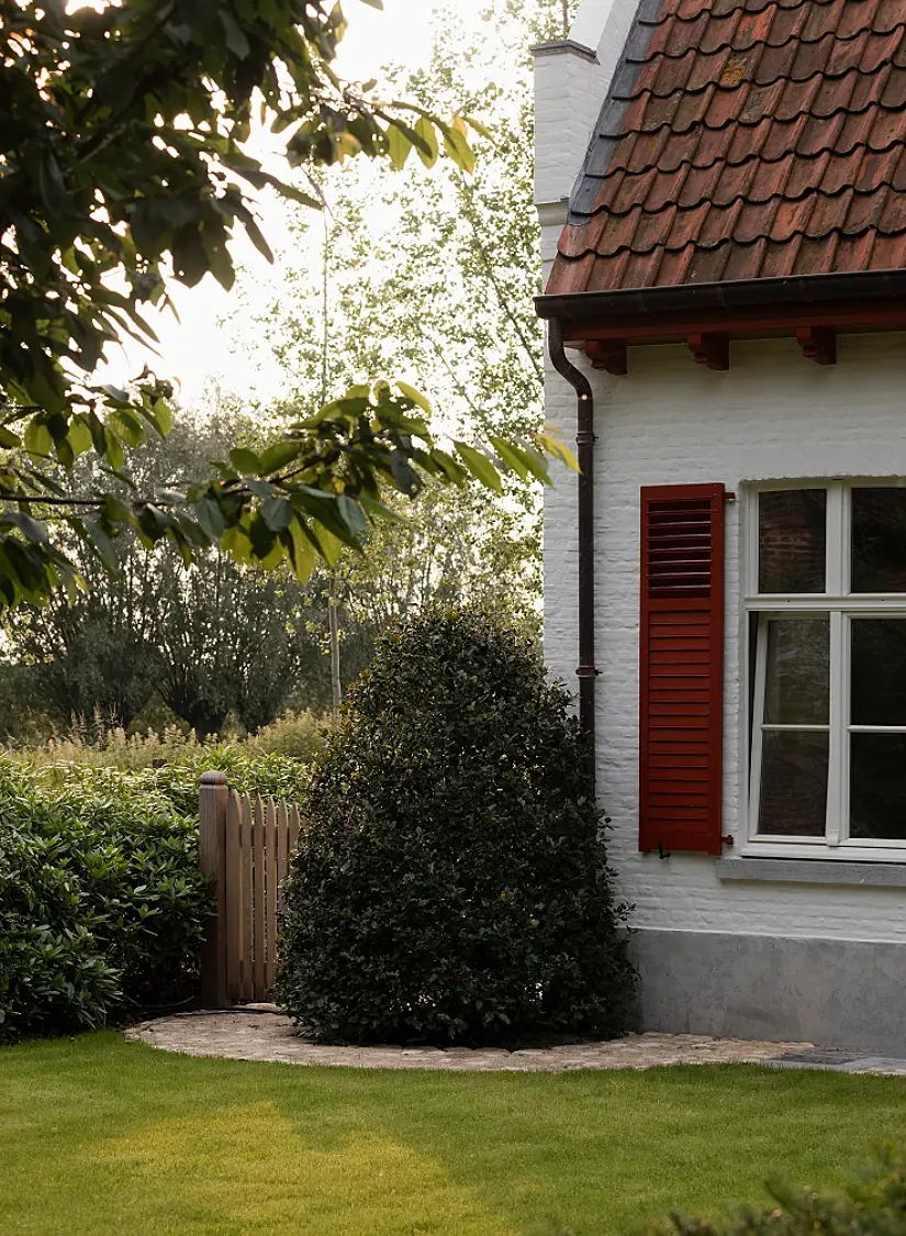 Witte gevel met rood luik, een kegel in groen, een houten poortje en een boord in kasseien
