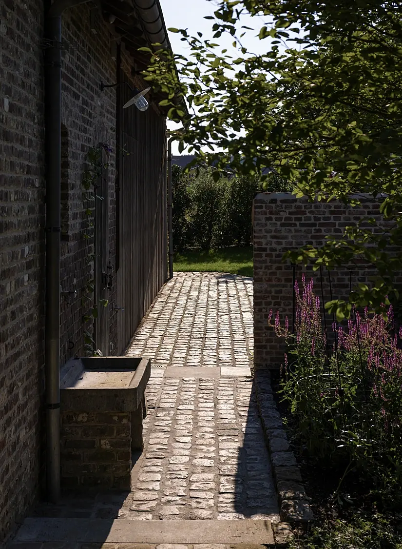 Smal pad in oude kasseien tussen bakstenen muren, met een stenen trog en vaste planten