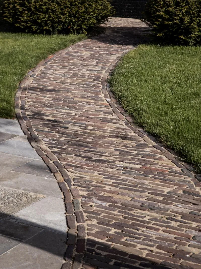 Gebogen pad in oude kasseien tussen gazon en geschoren taxus, met platen in grijze natuursteen