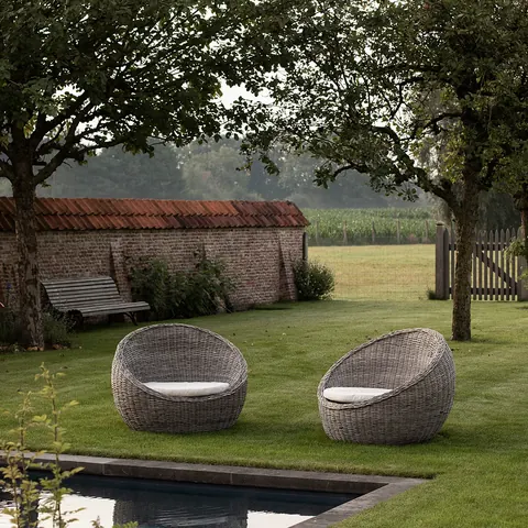 Twee rieten stoelen op het gazon onder fruitbomen, met een oude tuinmuur