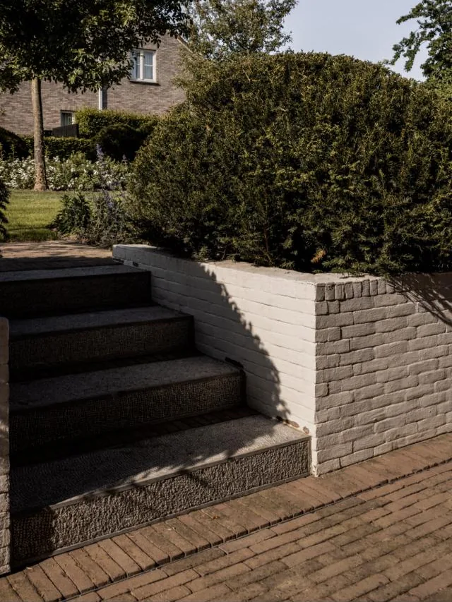 Trap in natuursteen tussen wit geschilderde muurtjes, met een pad in klinkers en een geschoren taxus